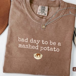 May include: Espresso-colored t-shirt with the text "bad day to be a mashed potato" in white. The shirt features a small graphic of mashed potatoes in a bowl. A silver necklace with a pendant is on the shirt.