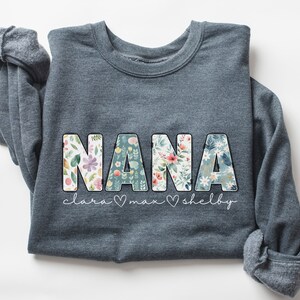 以下が含まれることがあります： 花柄の文字で「NANA」と書かれたグレーのスウェットシャツ。 「clara &hearts; max &hearts; shelby」という文字が「NANA」の下に印刷されています。