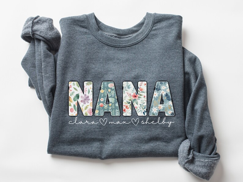 以下が含まれることがあります： 花柄の文字で「NANA」と書かれたグレーのスウェットシャツ。 「clara &hearts; max &hearts; shelby」という文字が「NANA」の下に印刷されています。