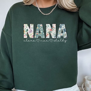 以下が含まれることがあります： 花柄の文字で「NANA」と書かれた濃い緑色のスウェットシャツ。 「NANA」の下には「clara &hearts; max &hearts; shelby」という文字が書かれています。