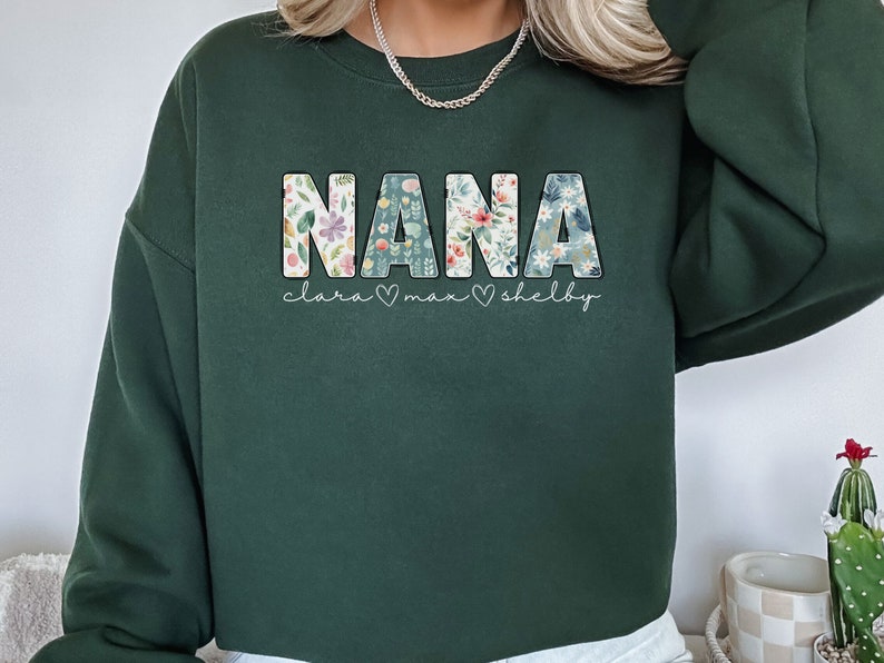 以下が含まれることがあります： 花柄の文字で「NANA」と書かれた濃い緑色のスウェットシャツ。 「NANA」の下には「clara &hearts; max &hearts; shelby」という文字が書かれています。