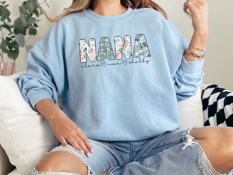 以下が含まれることがあります： 花柄のデザインが施された大きな文字で「NANA」と書かれた薄いブルーのトレーナー。文字は黒で縁取られており、その下に「Clara」、「Max」、「Shelby」という名前が小さなフォントで書かれている。