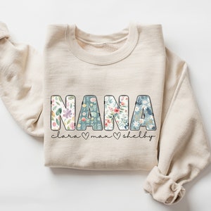 以下が含まれることがあります： ベージュのトレーナーに、大きな文字で「NANA」と書いてあります。文字は花柄で飾られています。「NANA」の下には「clara &hearts; max &hearts; shelby」という文字があります。