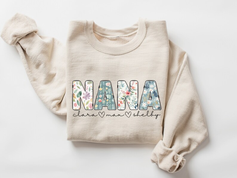 以下が含まれることがあります： ベージュのトレーナーに、大きな文字で「NANA」と書いてあります。文字は花柄で飾られています。「NANA」の下には「clara &hearts; max &hearts; shelby」という文字があります。