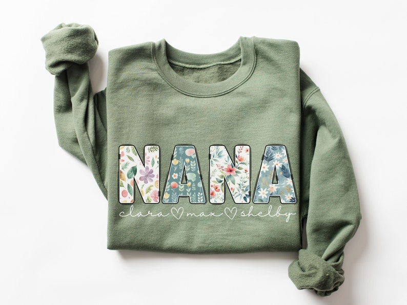 以下が含まれることがあります： 花柄の文字で「NANA」と書かれた緑色のスウェットシャツ。 「NANA」の下には、「Clara」、「Max」、「Shelby」という名前が筆記体で書かれています。