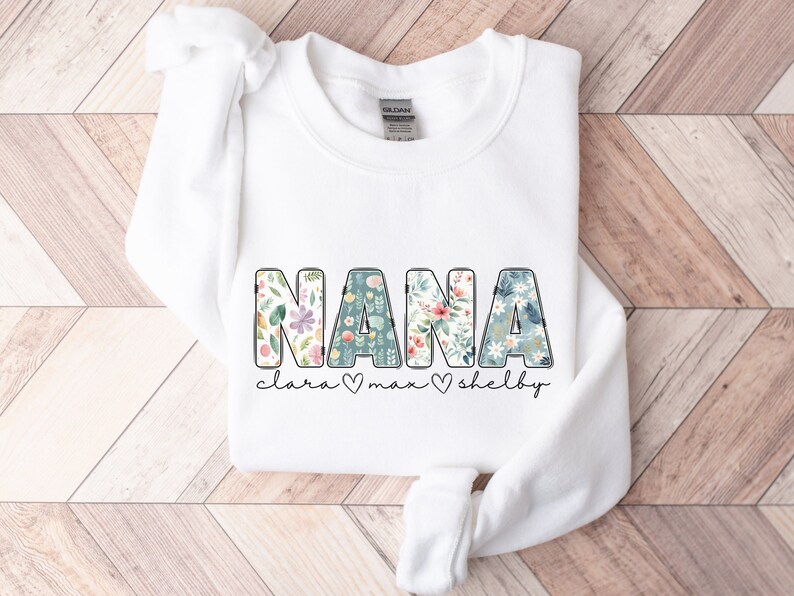 以下が含まれることがあります： 白いスウェットシャツに、花柄の文字で「NANA」と書かれています。「NANA」の下には、「Clara」、「Max」、「Shelby」という名前が、ハートマークで区切られて書かれています。