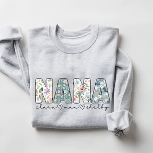 以下が含まれることがあります： 花のプリント文字で「NANA」と書かれたグレーのスウェットシャツ。 「NANA」の下には、「Clara」、「Max」、「Shelby」という名前がハートマークで区切られて書かれています。