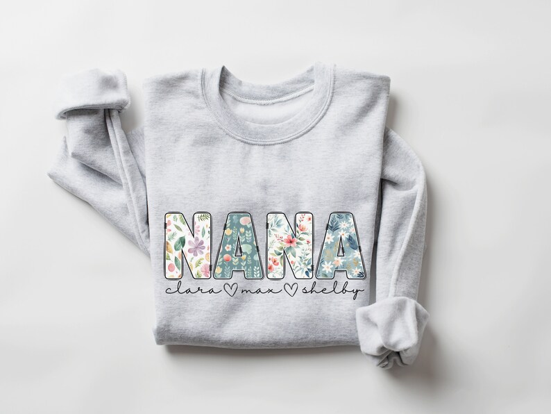 以下が含まれることがあります： 花のプリント文字で「NANA」と書かれたグレーのスウェットシャツ。 「NANA」の下には、「Clara」、「Max」、「Shelby」という名前がハートマークで区切られて書かれています。
