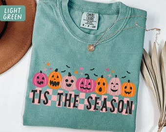 May include: Light green Comfort Colors t-shirt with a Halloween design. The shirt features a row of smiling pumpkins in pink and orange, with the text "Tis the Season" in a pink and black checkered pattern.