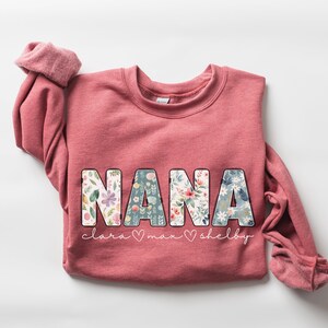 以下が含まれることがあります： 花柄の文字で「NANA」と書かれたピンク色のスウェットシャツ。 「NANA」の下には、「Clara」、「Max」、「Shelby」という名前がより小さなフォントで書かれている。