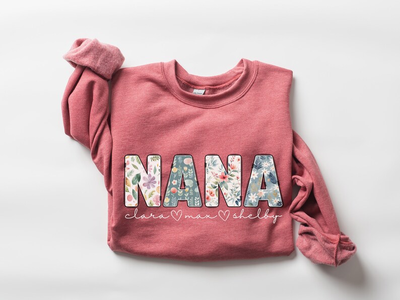 以下が含まれることがあります： 花柄の文字で「NANA」と書かれたピンク色のスウェットシャツ。 「NANA」の下には、「Clara」、「Max」、「Shelby」という名前がより小さなフォントで書かれている。