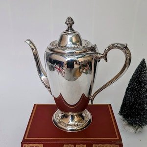 以下が含まれることがあります： 蓋に花柄の装飾が施された、装飾的な取っ手を持つ銀製のティーポット。ティーポットは、チョーサーの「カンタベリー物語」というタイトルの赤い本の上に置かれています。