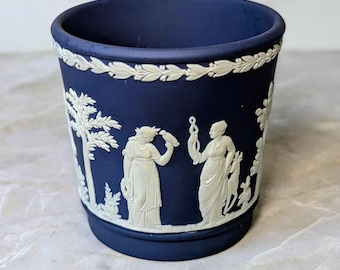 May include: A dark blue cylindrical ceramic vessel featuring white relief ornamentation. The exterior displays classical figures, trees, and foliage in a matte finish. A decorative laurel leaf border encircles the upper rim of the piece.