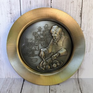 Op de afbeelding: Een goudkleurig metalen bord met een reliëfdesign van een man die een kalkoen met een mes snijdt. De scène speelt zich af aan een tafel met andere mensen om hem heen. Het bord heeft een verhoogde rand en een glad, glanzend oppervlak.