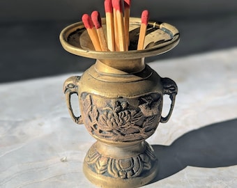 May include: A small, antique-style brass vessel with two side handles and a wide, flared rim. The body features embossed floral and bird motifs, with a pedestal base. Several wooden matches with red tips are placed upright inside the container.