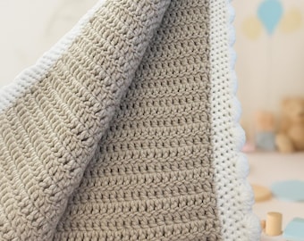 May include: A handmade crochet blanket in a neutral color palette. The blanket features a central section in a light tan color, bordered by a white scalloped edge. The soft texture and color scheme suggest a cozy, comforting item, perfect for a baby.