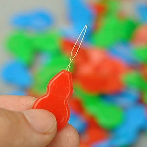 May include: A red plastic needle threader with a gold metal needle. The threader is shaped like a gourd.
