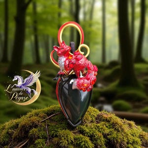 Puede incluir: Colgante de zorro rojo y dorado con base negra en forma de corazón. El zorro está detallado con gemas rojas y se asienta sobre el corazón. El colgante tiene un lazo para colgar. El fondo es una escena borrosa del bosque.