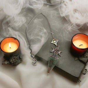 Puede incluir: Un péndulo plateado y verde con un diseño de triple luna y pentagrama cuelga de una cadena plateada. El péndulo está sobre una superficie gris con dos velas.