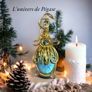May include: A gold wire wrapped pendant featuring a blue labradorite gemstone. The pendant is surrounded by a gold wire design and a small blue gemstone. The pendant is sitting on a white surface with a candle and pine branches in the background.