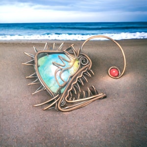 May include: A wire sculpture of a deep sea anglerfish with a blue and green iridescent glass eye. The sculpture is on a sandy beach with a red bead on the end of the wire.