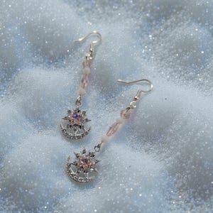 Peut inclure: Une paire de boucles d'oreilles argentées avec un motif céleste. Chaque boucle d'oreille présente un pendentif croissant de lune et étoile, orné de cristaux clairs et roses. Les boucles d'oreilles ont une chaîne de perles roses et blanches et une fermeture à crochet.
