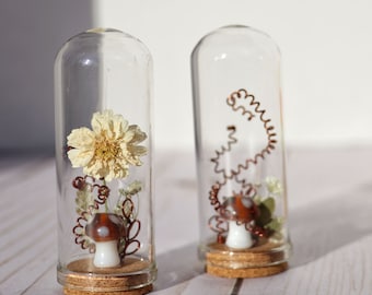 May include: Two glass bottles with cork stoppers, each containing a small mushroom and dried flowers. One bottle has a white flower and the other has a copper wire.