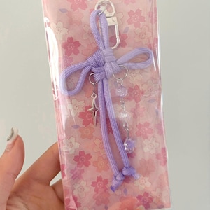 May include: A lavender-colored keychain with a silver clasp and decorative charms. The charms include a star and dangling beads. The keychain is packaged in a clear plastic sleeve with a pink floral pattern.