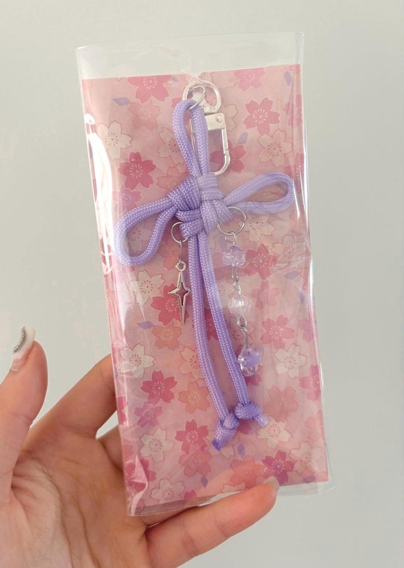 May include: A lavender-colored keychain with a silver clasp and decorative charms. The charms include a star and dangling beads. The keychain is packaged in a clear plastic sleeve with a pink floral pattern.