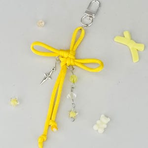 May include: A yellow keychain with a bow design, a silver-colored clasp, and dangling charms. The charms include a star-shaped pendant, clear and yellow beads, and a star-shaped charm. Additional yellow and white decorative elements are scattered around.