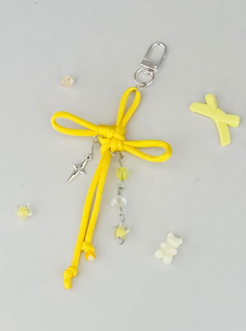 May include: A yellow keychain with a bow design, a silver-colored clasp, and dangling charms. The charms include a star-shaped pendant, clear and yellow beads, and a star-shaped charm. Additional yellow and white decorative elements are scattered around.