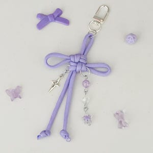 May include: A lavender keychain with a bow design, a silver star charm, and dangling beads. The keychain has a silver clasp and is surrounded by small decorative elements, including a bow, a butterfly, and a bead.