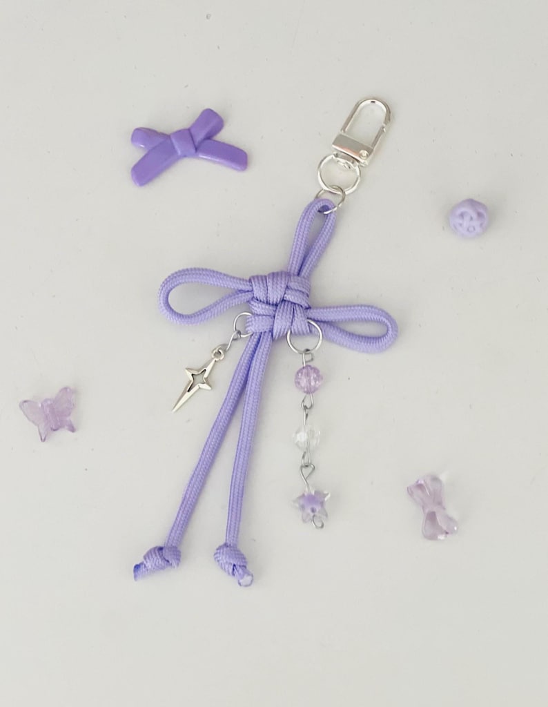 May include: A lavender keychain with a bow design, a silver star charm, and dangling beads. The keychain has a silver clasp and is surrounded by small decorative elements, including a bow, a butterfly, and a bead.