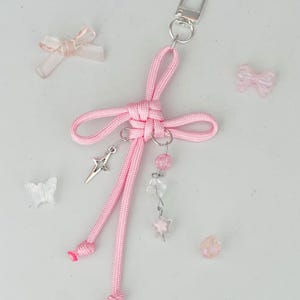 May include: A pink handmade keychain with a bow design, silver-tone hardware, and dangling charms. The charms include a star, pink beads, and clear beads. Several small, decorative bows and a butterfly are scattered around the keychain.