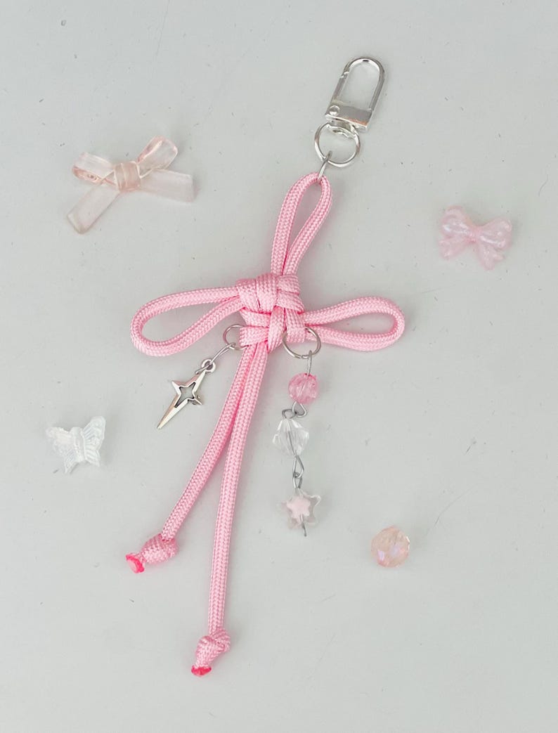 May include: A pink handmade keychain with a bow design, silver-tone hardware, and dangling charms. The charms include a star, pink beads, and clear beads. Several small, decorative bows and a butterfly are scattered around the keychain.