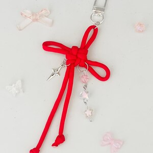May include: A red cord keychain with a silver clasp and decorative charms. The keychain features a knotted bow design, a silver star charm, and a strand of pink and clear beads. Additional small pink bows and beads are scattered around.