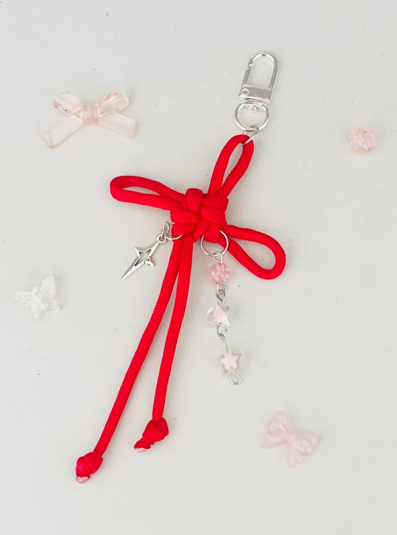 May include: A red cord keychain with a silver clasp and decorative charms. The keychain features a knotted bow design, a silver star charm, and a strand of pink and clear beads. Additional small pink bows and beads are scattered around.
