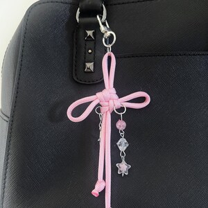 May include: A pink cord charm with a decorative knot and dangling beads and star charms. The charm is attached to a black handbag with silver hardware. The charm is a decorative accessory.