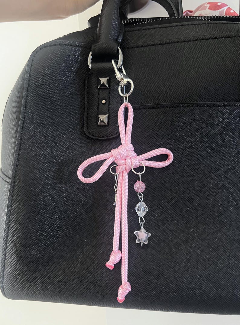 May include: A pink cord charm with a decorative knot and dangling beads and star charms. The charm is attached to a black handbag with silver hardware. The charm is a decorative accessory.