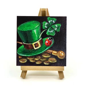 Può includere: Un dipinto in miniatura su un piccolo cavalletto di legno raffigura un cappello da folletto verde con fibbia marrone, un quadrifoglio, una coccinella e monete d'oro su sfondo nero. Il dipinto ha un tema del giorno di San Patrizio.
