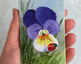 May include: A close-up of a painted canvas featuring a purple and white pansy flower with a ladybug on the petal. The flower is surrounded by green foliage.