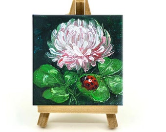 May include: A small painting on a wooden easel depicts a pink flower with green leaves and a red ladybug. The background is dark green.