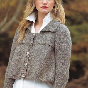 May include: A cropped, button-down, knitted jacket in a speckled grey colour. The jacket features a wide collar and is worn over a white collared shirt. The buttons are a light, neutral colour. The jacket is a classic style.