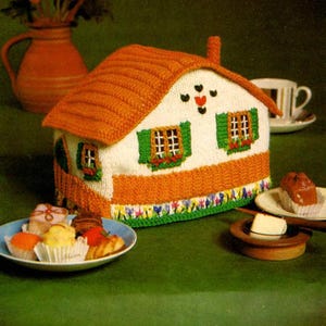 May include: A knitted teapot cozy shaped like a house with a brown roof, white walls, green shutters, and a flower garden at the base. The cozy is sitting on a green tablecloth with a plate of pastries and a cup and saucer.