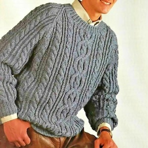 May include: A grey long-sleeve cable knit sweater with a crew neckline, worn over a light-colored collared shirt. The sweater features vertical textured cable patterns throughout the front and sleeves, paired with brown leather trousers.
