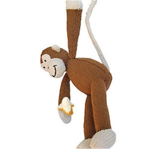 May include: A knitted monkey toy in brown and white, with a long tail. The monkey features a white face, hands, and feet, and holds a yellow and white banana. It is hanging upside down.