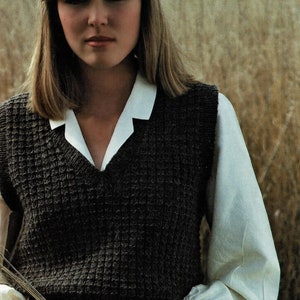 May include: A woman wearing a brown knitted waistcoat over a white collared shirt with puffed sleeves. She is standing in a field of long grass.