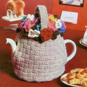 May include: A beige knitted teapot cozy with a colorful crocheted flower design. The cozy is on a red surface with a plate of pastries.