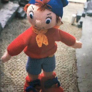 Può includere: Bambola fatta a mano all'uncinetto con cappello blu, maglione rosso, pantaloncini blu e scarpe rosse. La bambola ha una sciarpa gialla e capelli arancioni. La bambola è in piedi su una superficie di pietra.