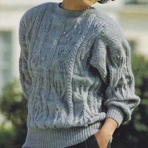 Könnte beinhalten: Ein grauer Strickpullover mit einem Zopfmuster. Der Pullover hat einen Rundhalsausschnitt und lange Ärmel.